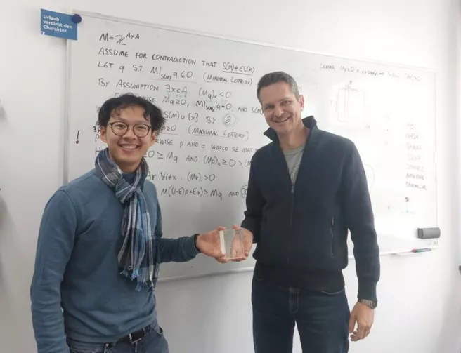 Chris Dong reicht dem Preisträger, Prof. Brandt, die Trophäe. Im Hintergrund ist ein vollgeschriebenes Whiteboard zu sehen.